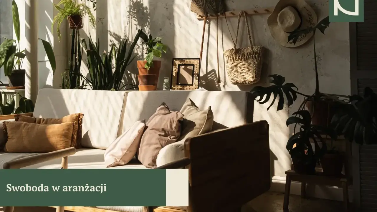 Przytulny salon boho z dużą ilością roślin, plecionym koszem i słomkowym kapeluszem na ścianie. Miękkie poduszki na kanapie zapraszają do relaksu.