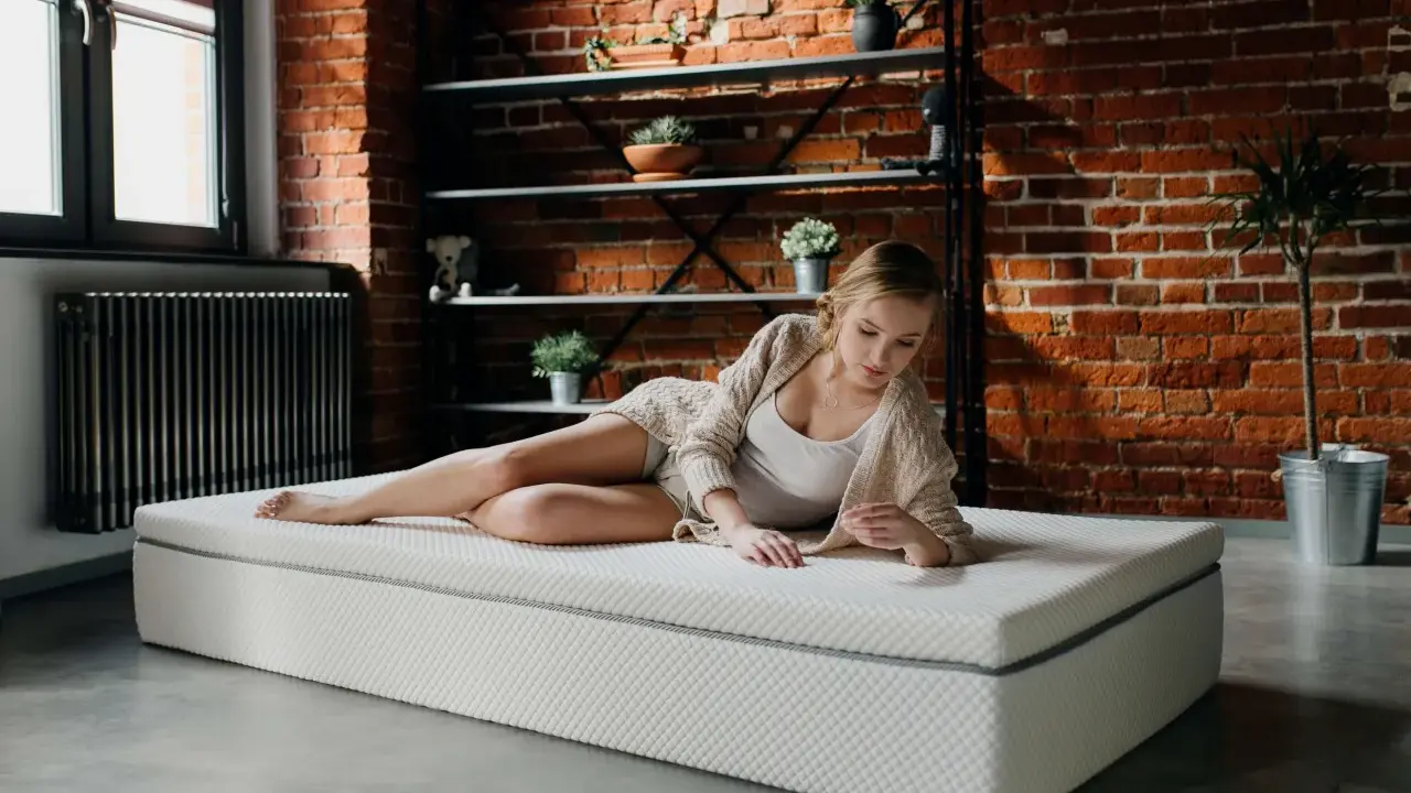 Młoda kobieta odpoczywa na materacu na ziemi w industrialnym wnętrzu. Ciemna ceglana ściana i metalowe półki tworzą tło.