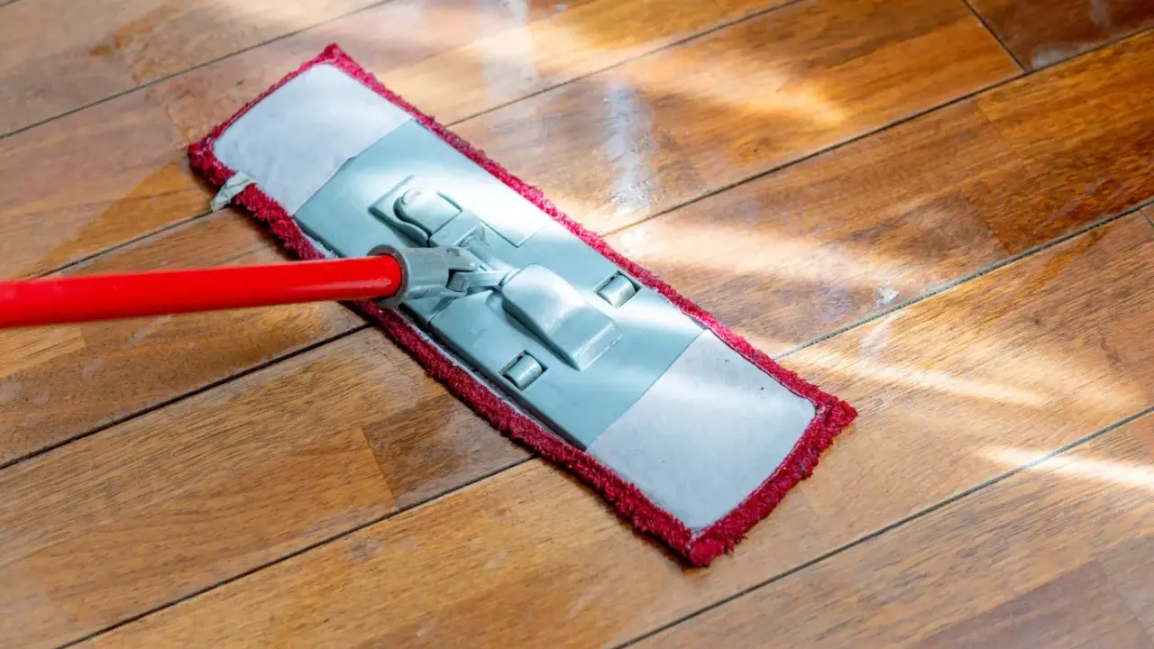 Czerwony mop z płaską końcówką czyści drewnianą podłogę. Słońce odbija się od mokrej powierzchni, pokazując, jak myć podłogę.