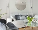Jasny salon z białymi ścianami i meblami. Miękka kanapa, szare fotele i okrągłe lustro tworzą przytulną atmosferę.