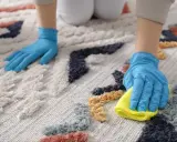 Ręce w niebieskich rękawiczkach czyszczą dywan. Jedna ręka rozsypuje sodę oczyszczoną, druga wyciera plamę żółtą ściereczką.