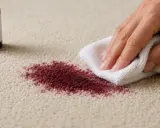 Ręka z białą ściereczką usuwa plamę z dywanu. Pranie dywanu płynem do naczyń to skuteczny sposób na pozbycie się zabrudzeń.