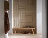 Nowoczesny pomysł na ścianę w korytarzu: tapicerowane panele w geometryczne wzory, drewniana ławka, płaszcz i buty.