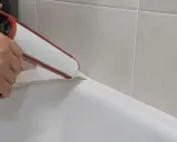 Ręka trzymająca pistolet do fugowania, nakładająca białą fugę na brzeg wanny, tworząc idealną linię.