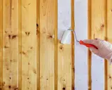 Ręka w rękawiczce maluje białą farbą drewniane panele ścienne. Proces malowania paneli ściennych papierowych trwa.