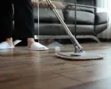 Osoba w kapciach czyści drewniany parkiet mopem. Proste sposoby, jak odnowić parkiet domowym sposobem, by znów lśnił.
