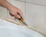 Ręka z nożem odcina stary silikon, pokazując, jak usunąć stary silikon z wanny.