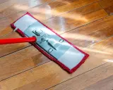 Czerwony mop z płaską końcówką czyści drewnianą podłogę. Słońce odbija się od mokrej powierzchni, pokazując, jak myć podłogę.