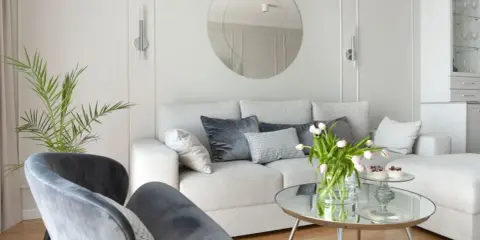 Jasny salon z białymi ścianami i meblami. Miękka kanapa, szare fotele i okrągłe lustro tworzą przytulną atmosferę.
