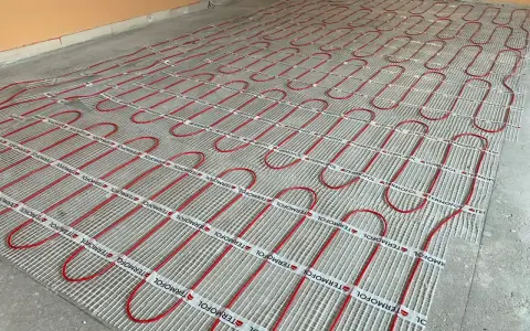 Instalacja ogrzewania podłogowego z czerwonymi rurkami na siatce. Kluczowe jest zachowanie odpowiedniej, minimalnej grubości wylewki cementowej dla efektywnego rozprowadzania ciepła.