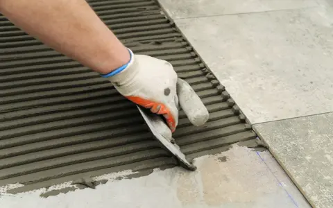 Ręka w rękawicy rozprowadza klej kielnią, przygotowując podłoże do tego, jak układać płytki na schodach zewnętrznych.