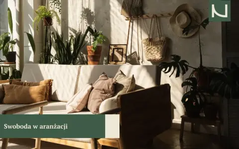 Przytulny salon boho z dużą ilością roślin, plecionym koszem i słomkowym kapeluszem na ścianie. Miękkie poduszki na kanapie zapraszają do relaksu.