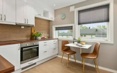 Mała kwadratowa kuchnia w bloku z oknem, z białymi szafkami, drewnianym blatem i dwoma brązowymi krzesłami przy małym stoliku.