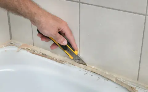 Ręka z nożem odcina stary silikon, pokazując, jak usunąć stary silikon z wanny.