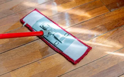Czerwony mop z płaską końcówką czyści drewnianą podłogę. Słońce odbija się od mokrej powierzchni, pokazując, jak myć podłogę.