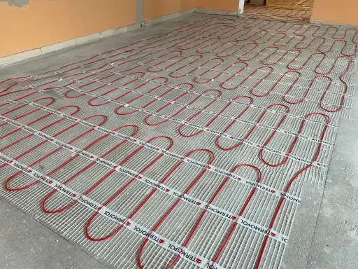 Instalacja ogrzewania podłogowego z czerwonymi rurkami na siatce. Kluczowe jest zachowanie odpowiedniej, minimalnej grubości wylewki cementowej dla efektywnego rozprowadzania ciepła.