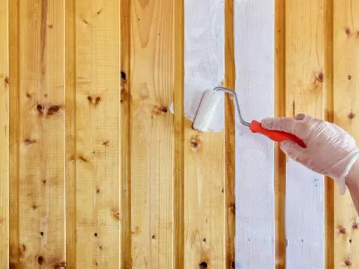 Ręka w rękawiczce maluje białą farbą drewniane panele ścienne. Proces malowania paneli ściennych papierowych trwa.