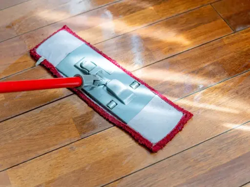 Czerwony mop z płaską końcówką czyści drewnianą podłogę. Słońce odbija się od mokrej powierzchni, pokazując, jak myć podłogę.