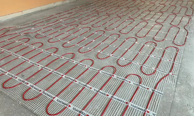 Instalacja ogrzewania podłogowego z czerwonymi rurkami na siatce. Kluczowe jest zachowanie odpowiedniej, minimalnej grubości wylewki cementowej dla efektywnego rozprowadzania ciepła.