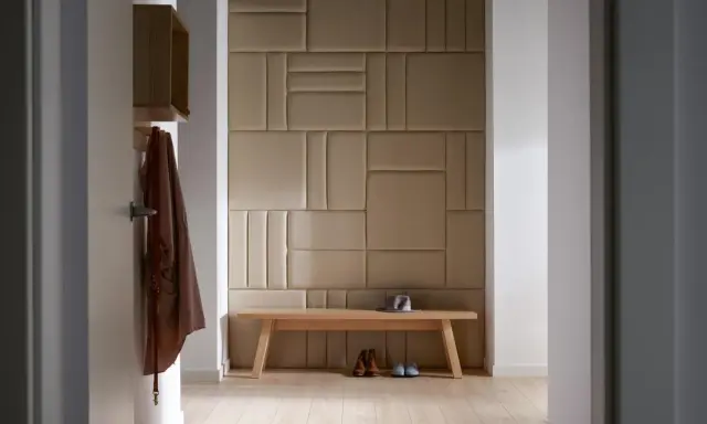 Nowoczesny pomysł na ścianę w korytarzu: tapicerowane panele w geometryczne wzory, drewniana ławka, płaszcz i buty.