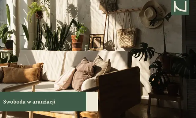 Przytulny salon boho z dużą ilością roślin, plecionym koszem i słomkowym kapeluszem na ścianie. Miękkie poduszki na kanapie zapraszają do relaksu.