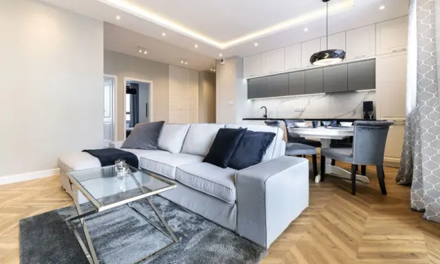 Nowoczesny salon z szarą sofą i szklanym stolikiem. Inspiracja, jak pomalować salon nowocześnie, z wykorzystaniem jasnych ścian i subtelnych dodatków.