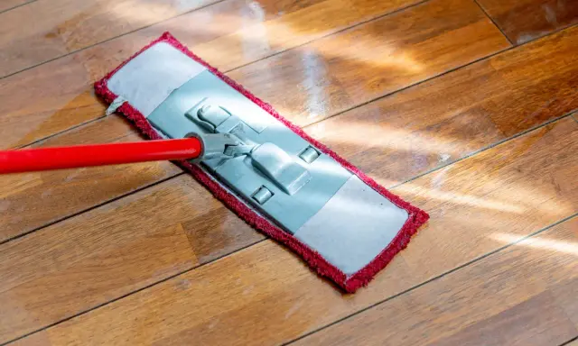 Czerwony mop z płaską końcówką czyści drewnianą podłogę. Słońce odbija się od mokrej powierzchni, pokazując, jak myć podłogę.