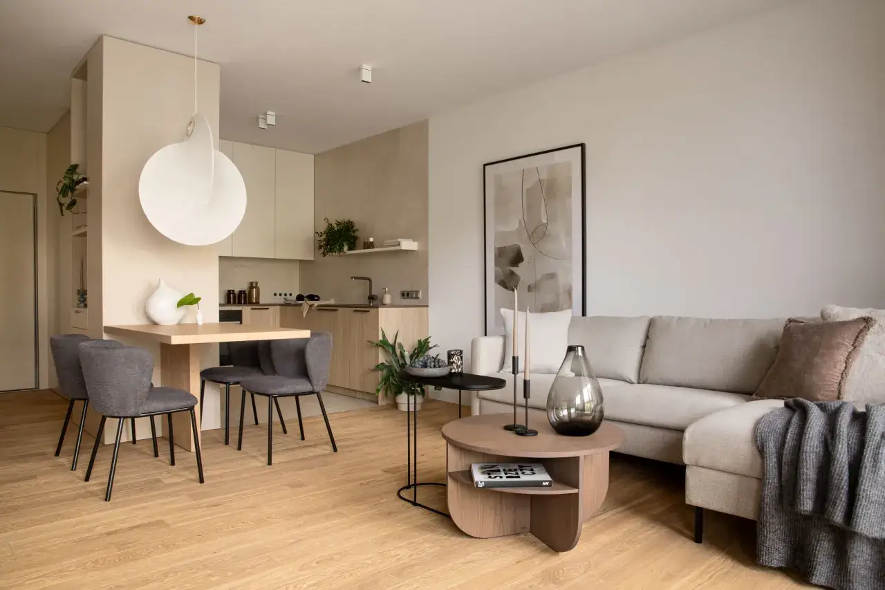 Przytulny salon w kolorach beżu, z wygodną kanapą, drewnianym stołem i nowoczesną kuchnią w tle.