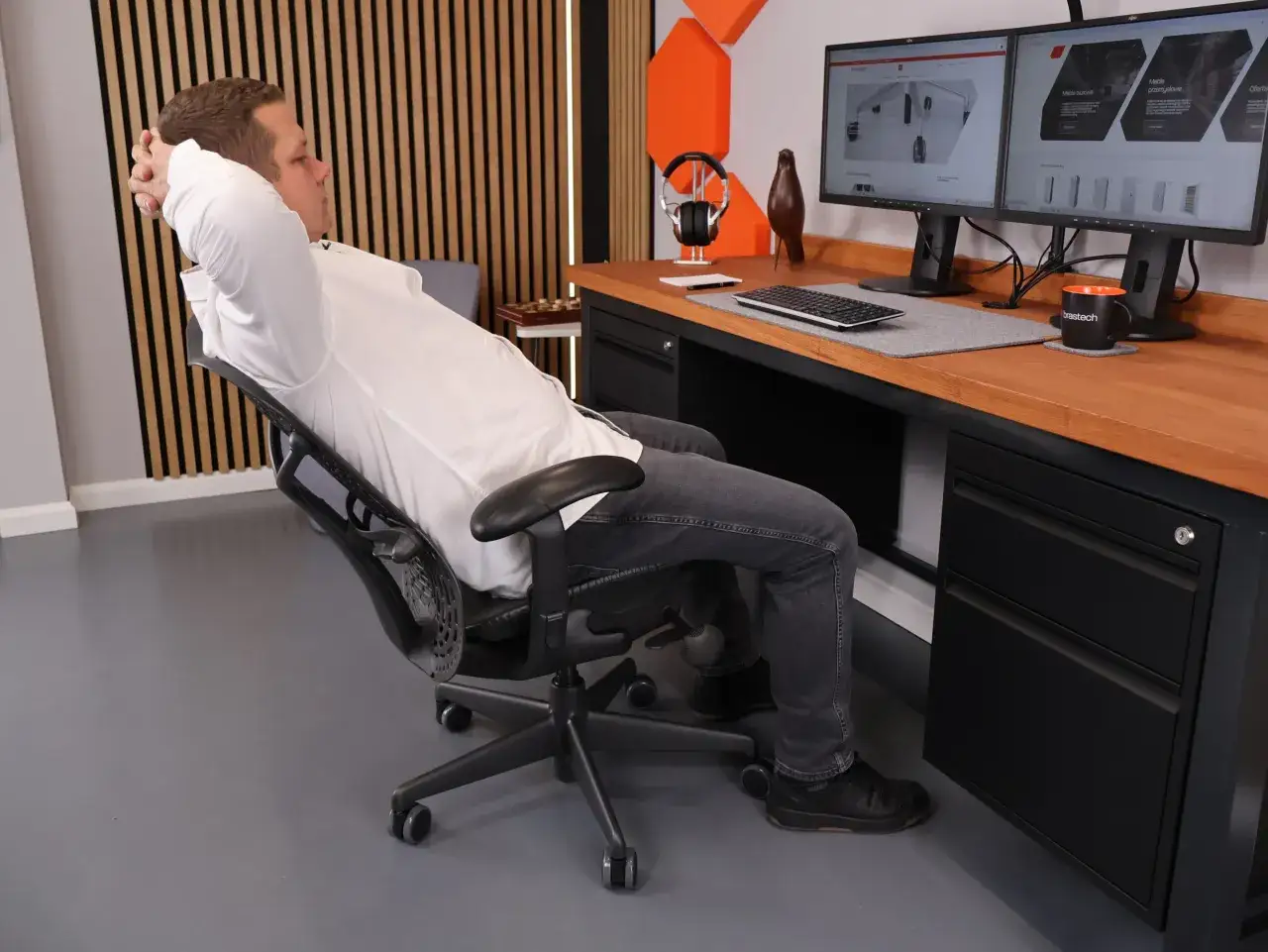 Mężczyzna odpoczywa przy biurku, opierając głowę na dłoniach. Widoczne są dwa monitory, klawiatura i kubek z logo. Ergonomiczna wysokość biurka zapewnia komfort.