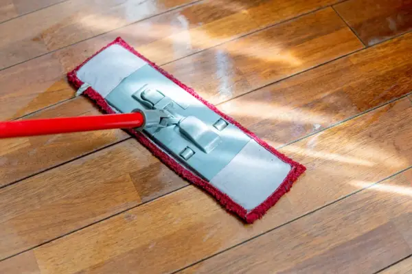 Czerwony mop z płaską końcówką czyści drewnianą podłogę. Słońce odbija się od mokrej powierzchni, pokazując, jak myć podłogę.